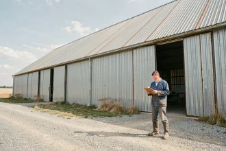 Agriculteur examinant des documents devant un grand hangar agricole en acier galvanisé dans une plaine rurale française